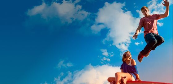 Deux personnes sur le toit d’un véhicule sous un ciel bleu, dont une en train de sauter.