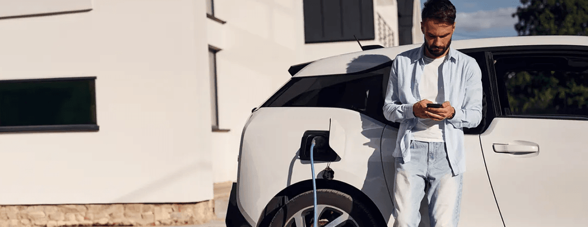 Personne utilisant un smartphone à côté d’une voiture électrique blanche en cours de recharge devant une habitation.
