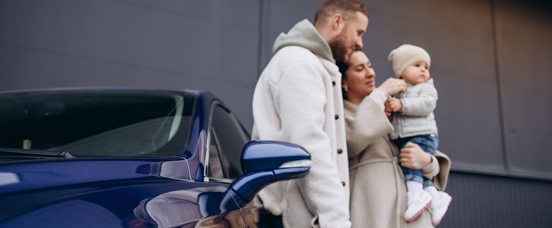 Personnes près d’une voiture bleue, dont un adulte tenant un bébé, devant un bâtiment.