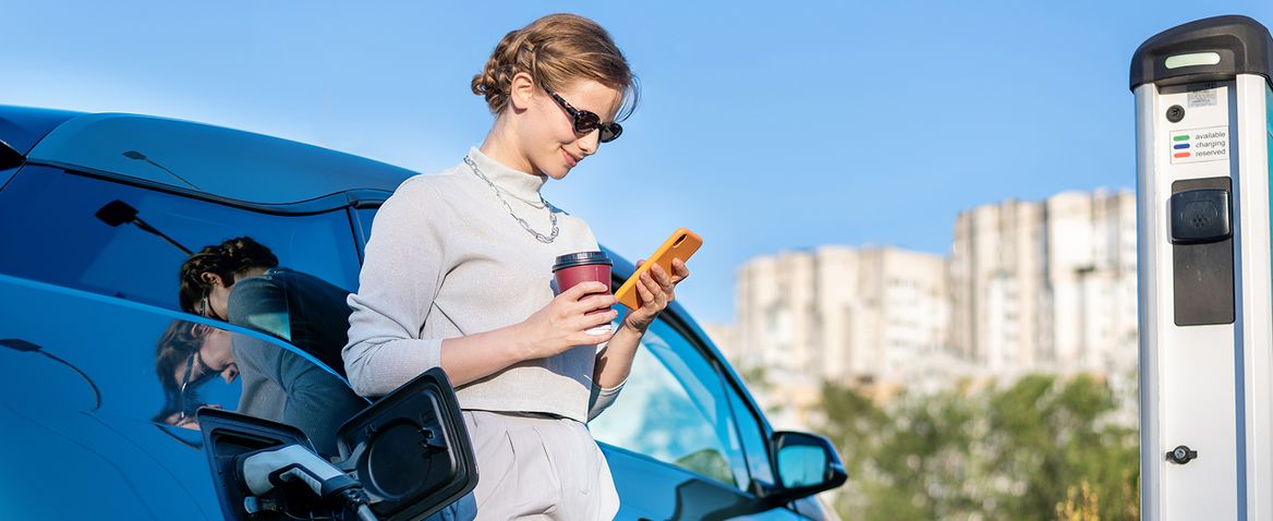 Personne utilisant un smartphone près d’une voiture électrique en cours de recharge à une borne extérieure.