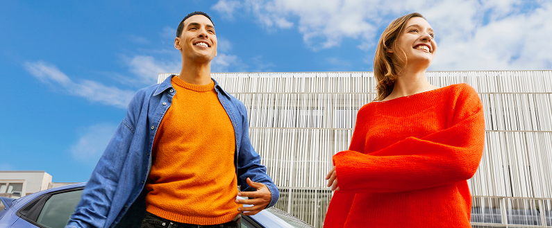 Deux personnes debout à côté d’un véhicule, portant des vêtements colorés, devant un bâtiment moderne à façade claire sous un ciel bleu.