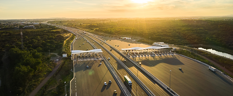 Vue aérienne d’une autoroute avec une barrière de péage au coucher du soleil, entourée de paysages verdoyants.